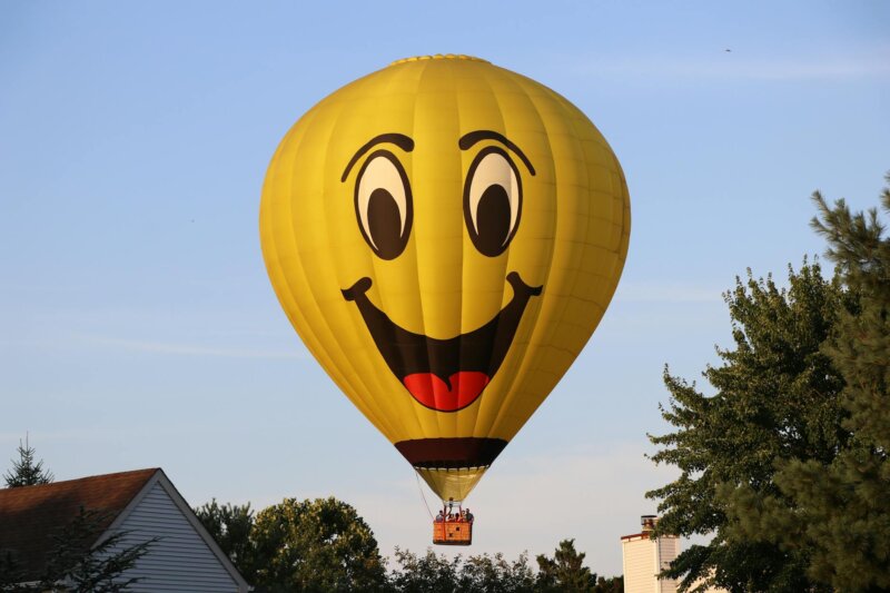 Bright yellow hot air balloon with a smiling face soaring over residential rooftops on a sunny day.