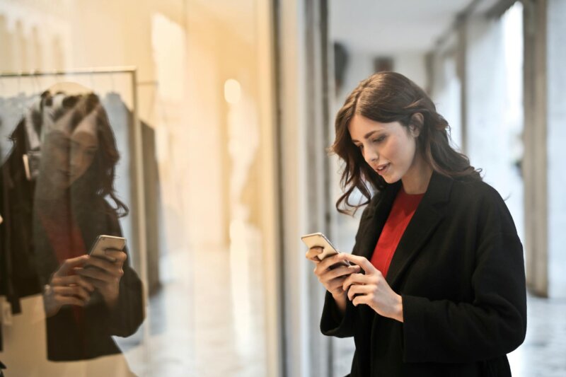 Young woman using smartphone at a store window reflecting her image, in casual fashion attire.