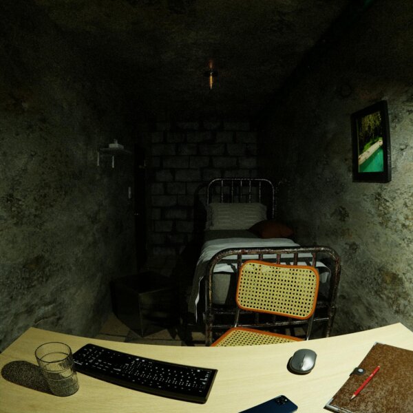 A dimly lit basement room with a computer setup and a simple bed frame, creating a rustic atmosphere.