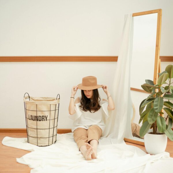 Caucasian woman sitting indoors with a hat, next to a laundry basket and mirror.